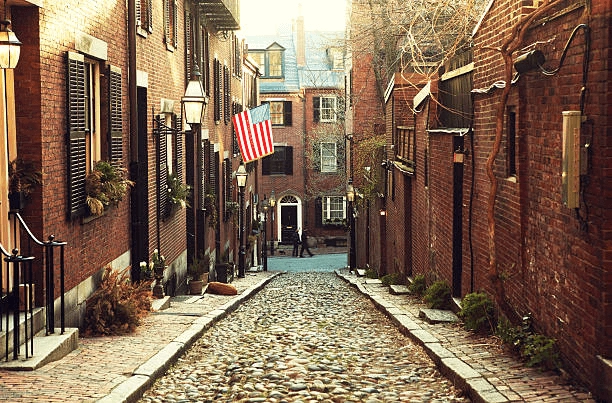 Cobblestone lane lined with brick buildings, lanterns and an American flag.