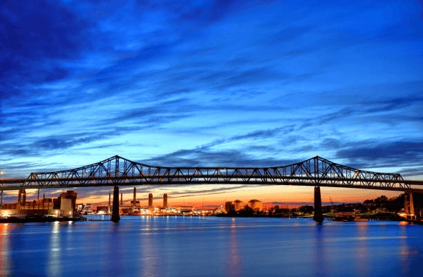 Steel-truss bridge crossing water at dusk, with an industrial shoreline beyond.