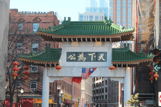 Green-roofed gateway with stone pillars, lanterns and flags above a city street.