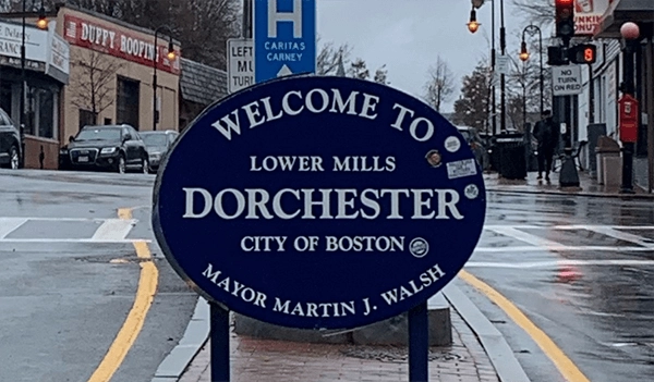 Welcome to Lower Mills Dorchester sign beside a wet street intersection.
