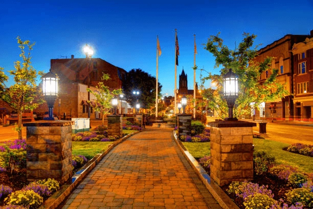 Brick walkway through a landscaped plaza with lanterns, flags and nearby buildings at dusk.
