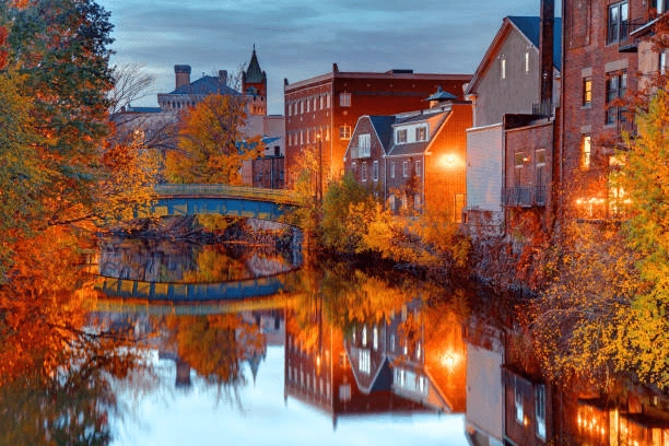 Brick buildings and autumn trees reflected in water beside a small bridge.