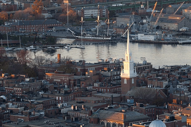 Brick rooftops and a church spire overlooking water and moored ships.