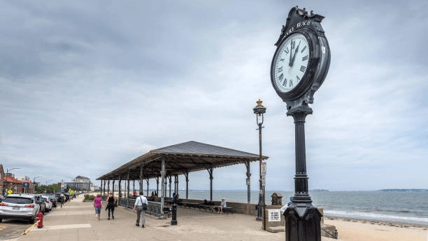 Seafront promenade with a street clock, shelter and beach beyond.