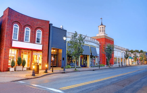 Low-rise storefronts and street lamps beside a road at dusk