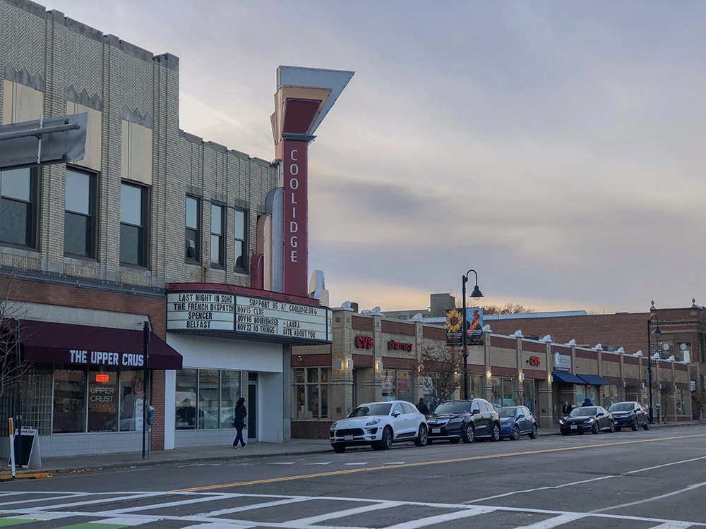 Cinema marquee and storefronts beside a street with parked cars and a crosswalk