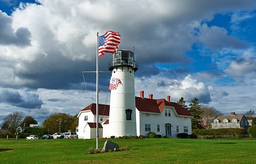 White lighthouse and red-roofed building beside a lawn and American flag