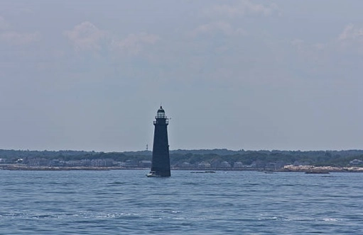 Dark lighthouse tower in open water with a low rocky shore behind it