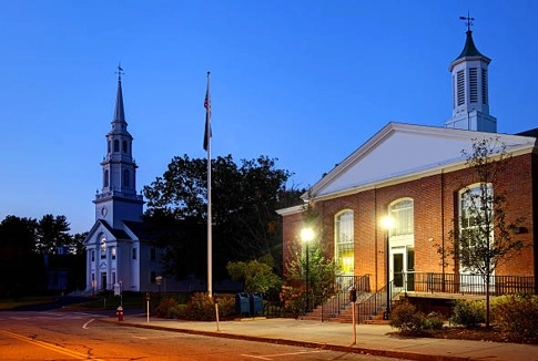 White steeple and brick building with a cupola beside a street at blue hour
