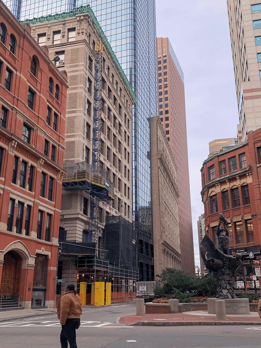 Brick, stone and glass buildings beside a city street with a statue and construction scaffolding