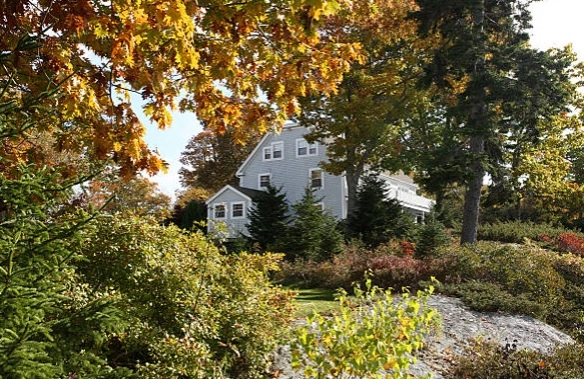 Gray house behind a garden with shrubs, a large boulder and autumn trees