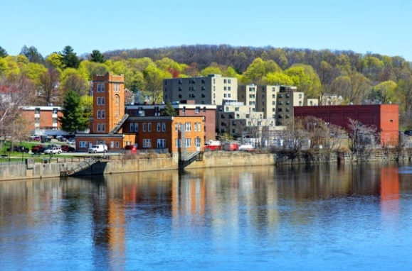 Brick and mid-rise buildings beside water with a wooded hillside behind them