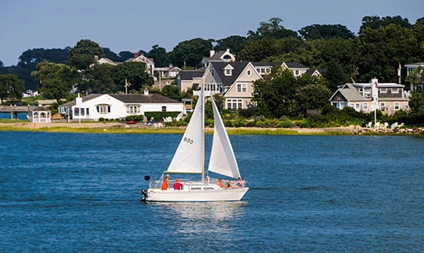 White sailboat on blue water with houses and trees along the shore