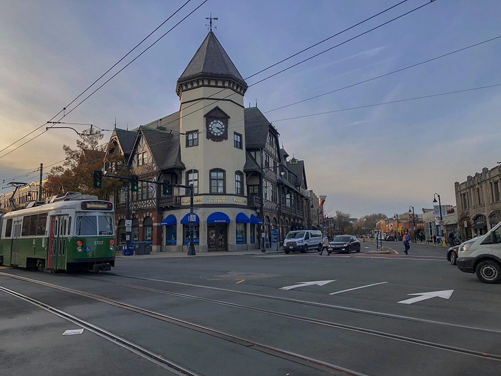 Clock-tower building, storefronts and a green railcar at a street crossing.