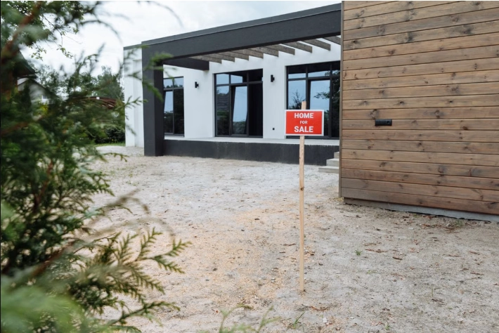 A red Home For Sale yard sign staked in the gravel driveway of a modern house