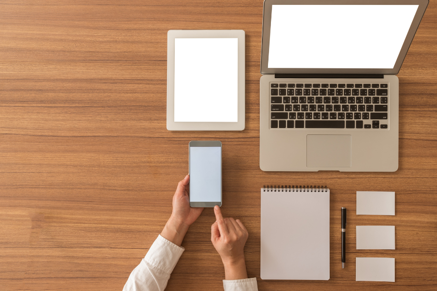 Photo of a laptop on wood desk with phone notepad and tablet with black pen and three white notecards