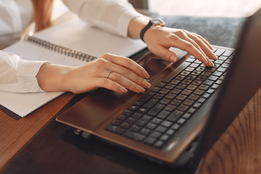 Typing on a laptop with an open notebook on a wood table